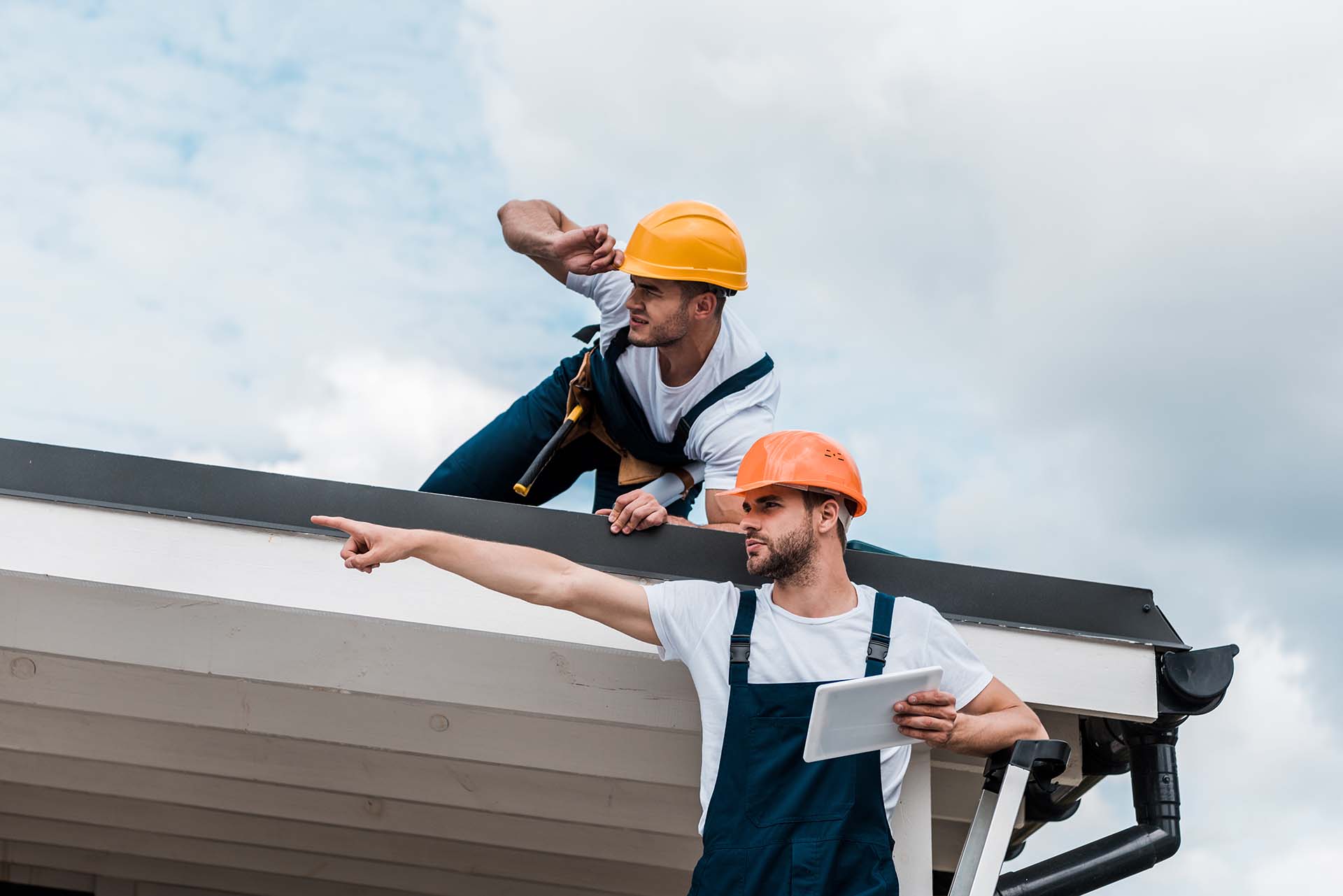 handsome bearded handyman in helmet holding digital tablet and pointing with finger near coworker